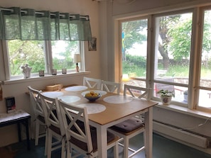 Uma sala de jantar com mesa posta para quatro pessoas, cadeiras brancas, um recipiente com limões e uma janela com vista para vegetação.