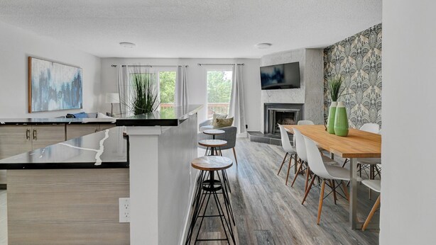A modern kitchen with a central island, bar stools, a dining area with a wooden table and chairs, a fireplace, and a TV mounted on the wall. - Skyline Views, Sleeps 8 and only Steps to SAIT, U of C & Downtown! (Calgary)