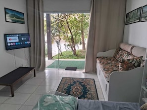Ein Wohnzimmer mit einer Couch, einer Bank, einem Fernseher und Blick auf einen Garten durch eine Glastür.