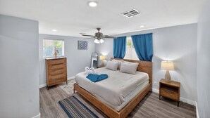 Quarto com cama, mesa de cabeceira, cômoda, ventilador de teto e janela com cortinas.