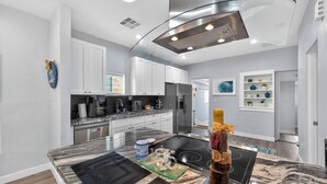 Uma cozinha moderna com armários brancos, uma ilha de vidro, eletrodomésticos de aço inoxidável e um design de teto diferenciado.