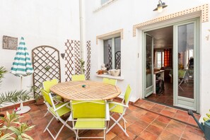 A patio with a round wooden table and four chairs, a striped umbrella, and a door leading to an interior space. - Patio Voltaire - A 5km de la plage (Sérignan)