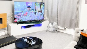 A modern living room with a flat-screen TV, a robotic vacuum cleaner, a standing fan, and a white sofa with a black cushion. - Welcome to Peace Pad 
Designed with comfort and convenience in mind. (Derby)