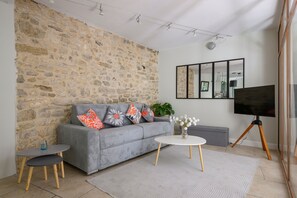 Un salón con pared de piedra, sofá gris, una mesa de centro redonda y un televisor sobre un trípode.