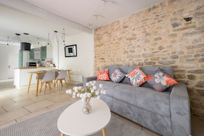 A modern living room with a grey sofa, a dining area with a wooden table, and a kitchen in the background.