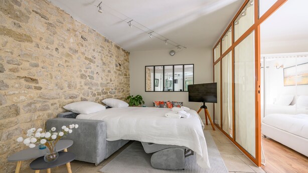 Ein Schlafzimmer mit Stehwand, einem Bett mit weißer Bettwäsche, einem kleinen Tisch mit einer Vase und einem Fernseher auf einem Schrank.