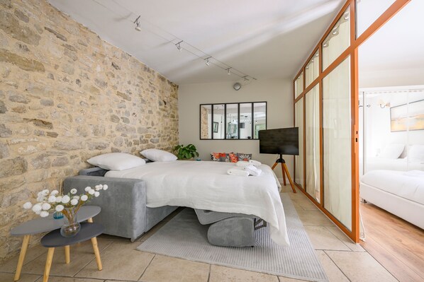 A bedroom with a stone wall, a bed with white bedding, a small table with a vase, and a TV on a stand.