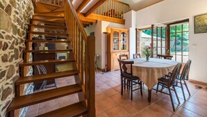 Una escalera de madera conduce a un segundo piso, un comedor con una mesa redonda y sillas, y vistas a través de amplios ventanales.