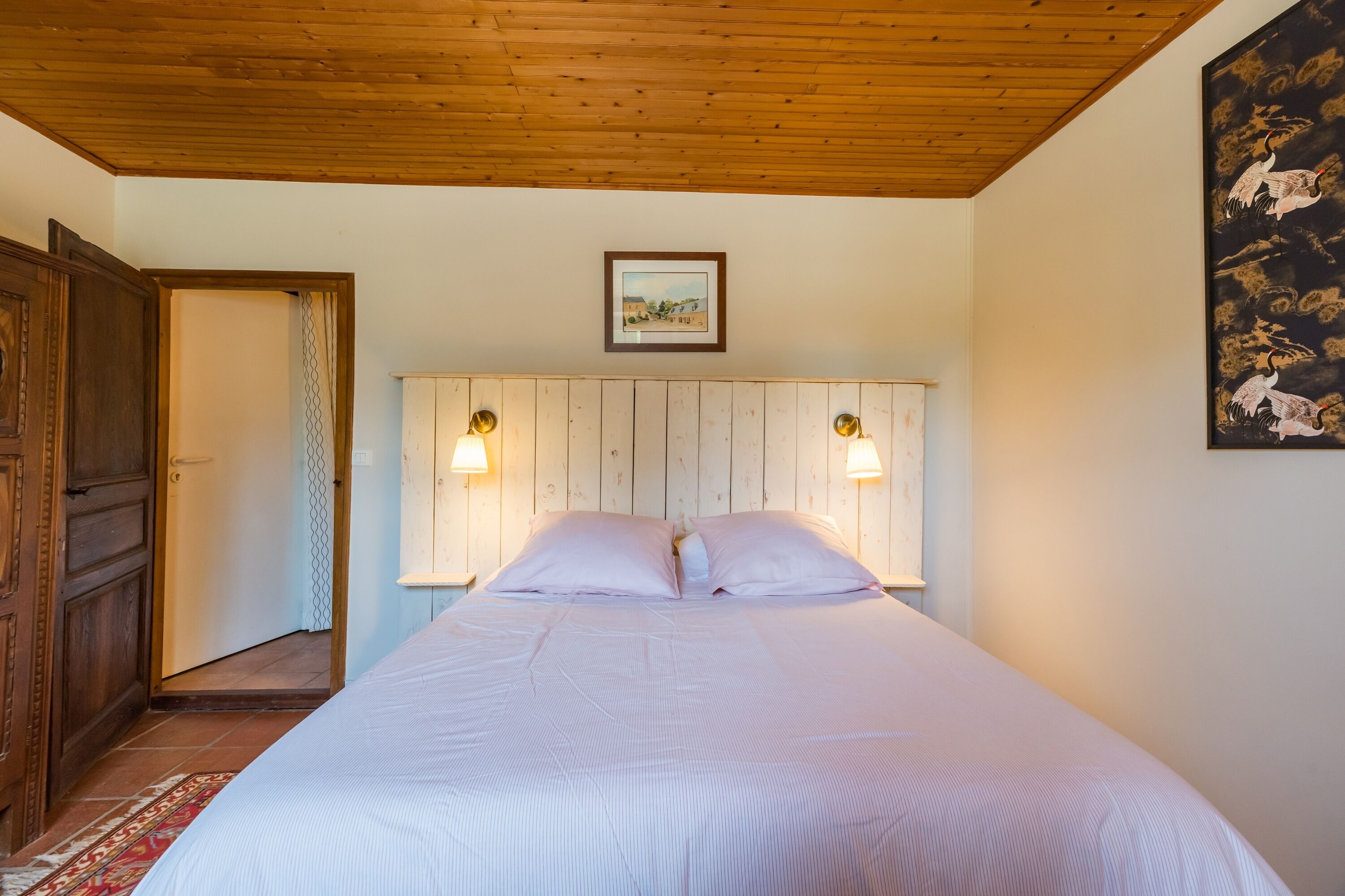 Une chambre avec un plafond en bois, un lit avec une literie blanche, deux lampes fixées au mur et un tableau encadré au-dessus du lit.