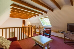 Ein gemütliches Dachzimmer mit Holzbalkendecke, einem kleinen Tisch, einem Stuhl, einer Couch, einem Fernseher und einem Fenster.