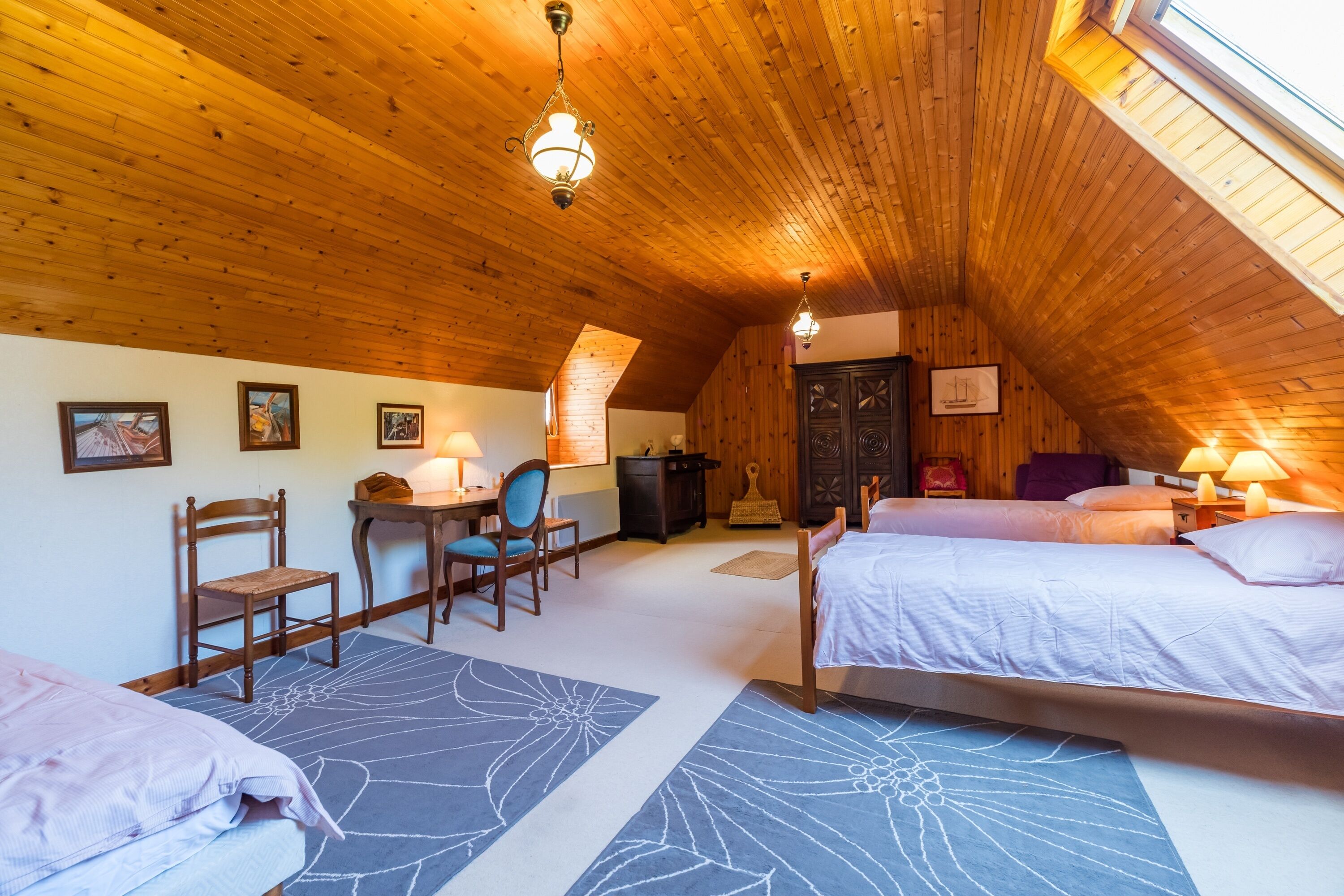 An attic bedroom with wooden ceiling, two beds, a desk, chairs, and a fireplace.