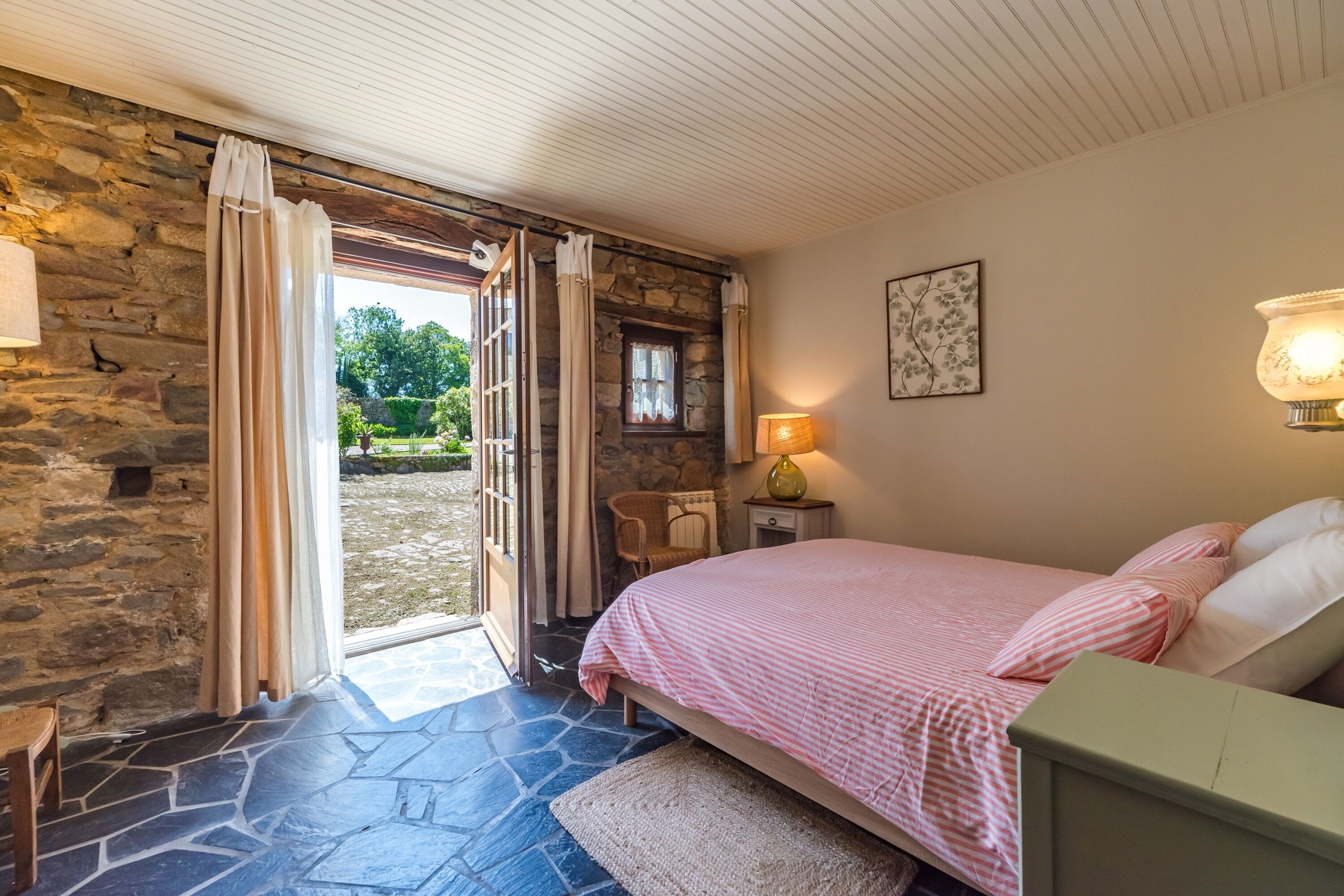 Un dormitorio con pared de piedra, una cama con ropa de cama rosa, una mesita de noche con lámpara y vistas al exterior.