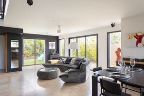 A spacious living area with a grey sectional sofa, a round ottoman, and a dining table set for four. Large windows provide a view of the outdoors. - La Villa Meunier - Four Bedroom House, Sleeps 7 (Dinard)