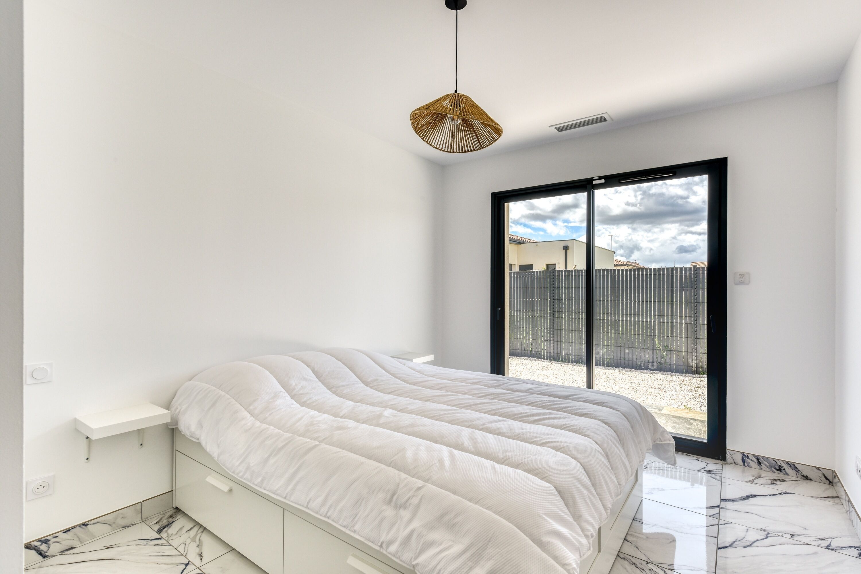 Un dormitorio con cama, lámpara colgante, puerta corrediza de vidrio y pared blanca.