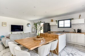 A modern kitchen with white cabinetry, a wooden dining table, and a built-in island. - L'Instant Minervois - Avec piscine privée (Olonzac)