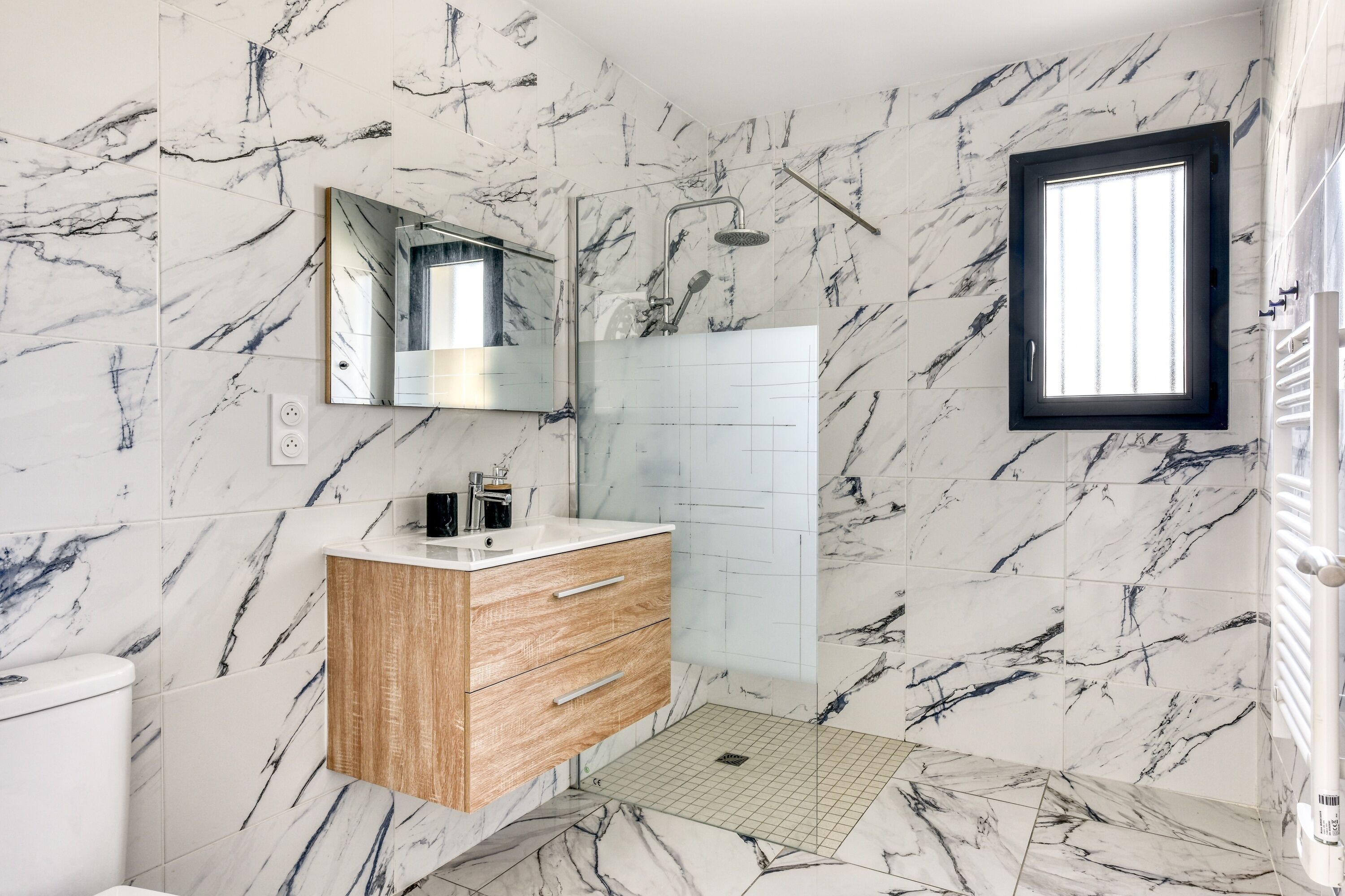 Une salle de bain avec des murs en marbre, un meuble-lavabo en bois, un miroir, une douche et une fenêtre.