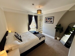 Ein Schlafzimmer mit Bett, Nachttischlampe, einem Fenster mit Vorhängen, einer Heizung, einem Wandregal, einem gerahmten Bild, einem Topf mit Pflanze und einem Spiegel.