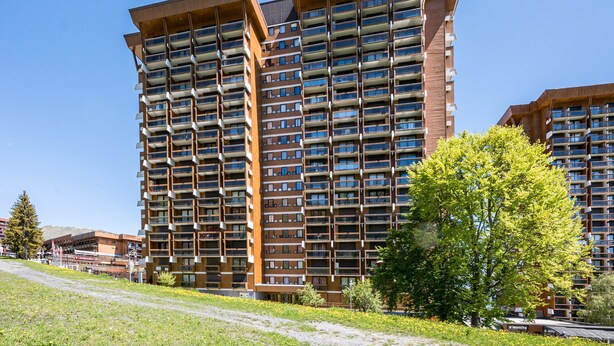 A high-rise residential building with multiple balconies, a clear sky, and a grassy area with trees. - Le Zodiaque - Station de ski accessible à pied (Villarembert)