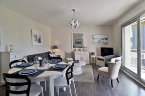 A dining area with a round table, chairs, and a chandelier. There is a sofa and a television in the background. - L'Échappée Dinardaise - À 400 m de la plage (Dinard)