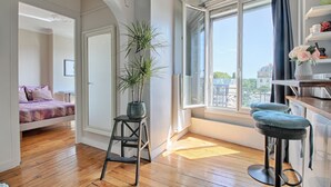 Una stanza con un letto, uno sgabello, una finestra e vista sugli edifici.