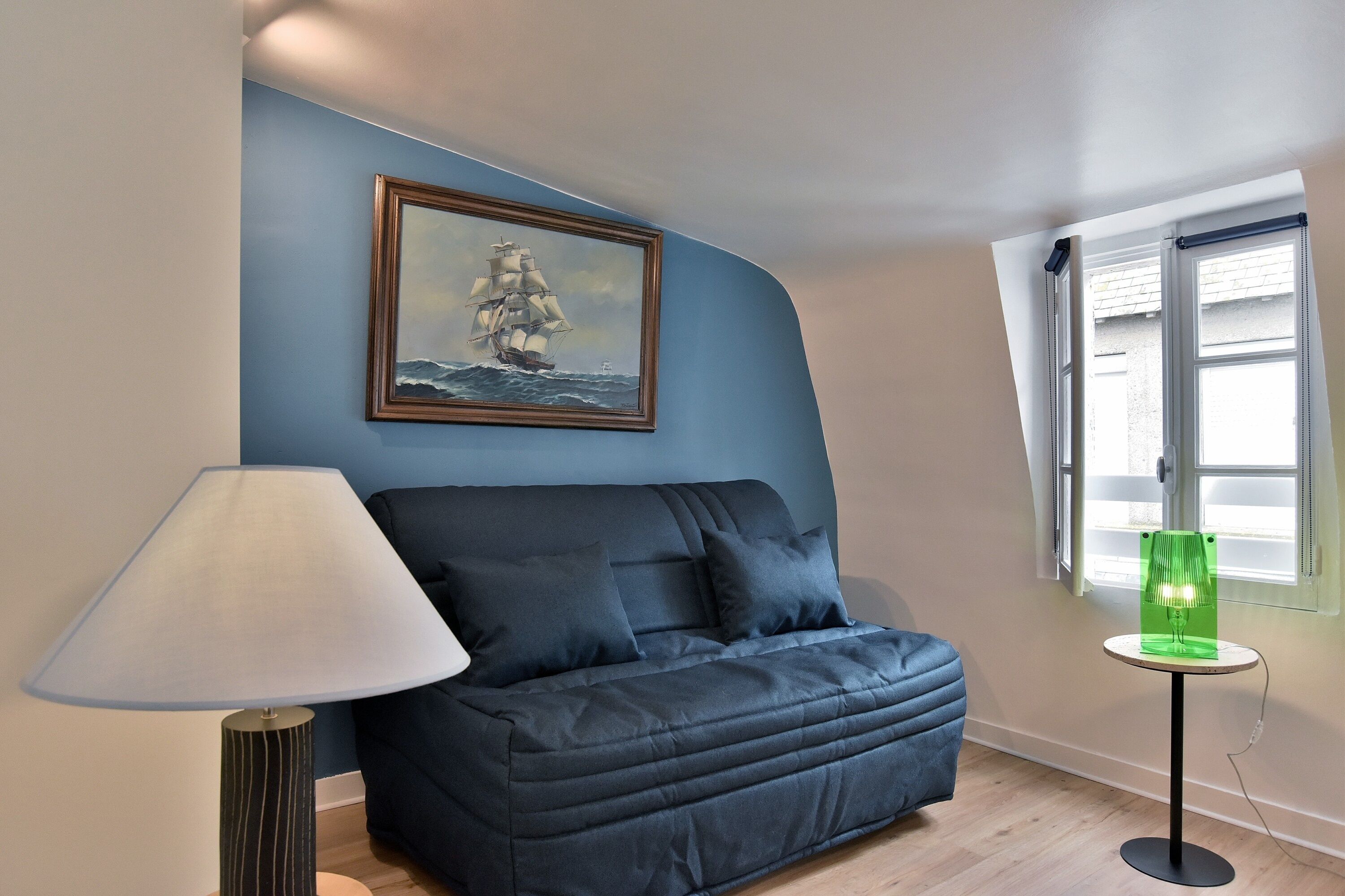 Una habitación con pared azul, un cuadro enmarcado de un barco, un sofá azul oscuro, una lámpara y una lámpara de mesa verde.
