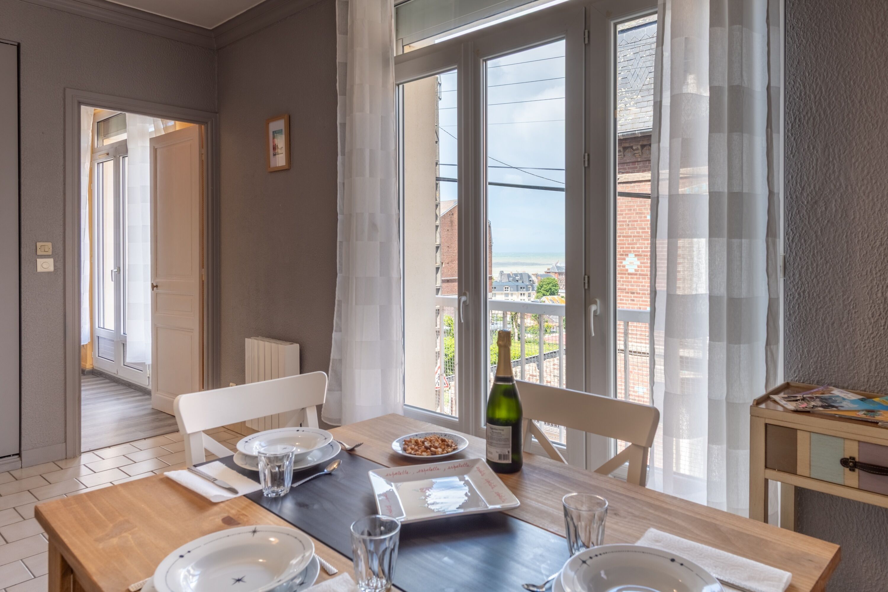 Un coin repas avec une table dressée pour deux personnes, une bouteille de vin et une vue sur la mer grâce à de grandes fenêtres.