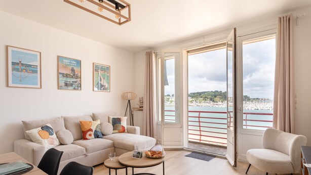 A modern living room with a sofa, coffee table, and chairs, a balcony with a railing, and a cityscape view. - Bel Horizon Marin - Magnifique vue mer (Saint-Malo)
