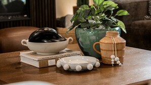 Una sala de estar con una mesa de centro de madera, una planta verde, un tazón blanco, un libro y una lámpara.