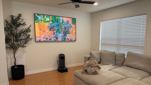 A living room with a beige sofa, a potted plant, a colorful abstract painting, a ceiling fan, and a window with blinds. - 3 Bedroom Home with Private Pool (Schertz)