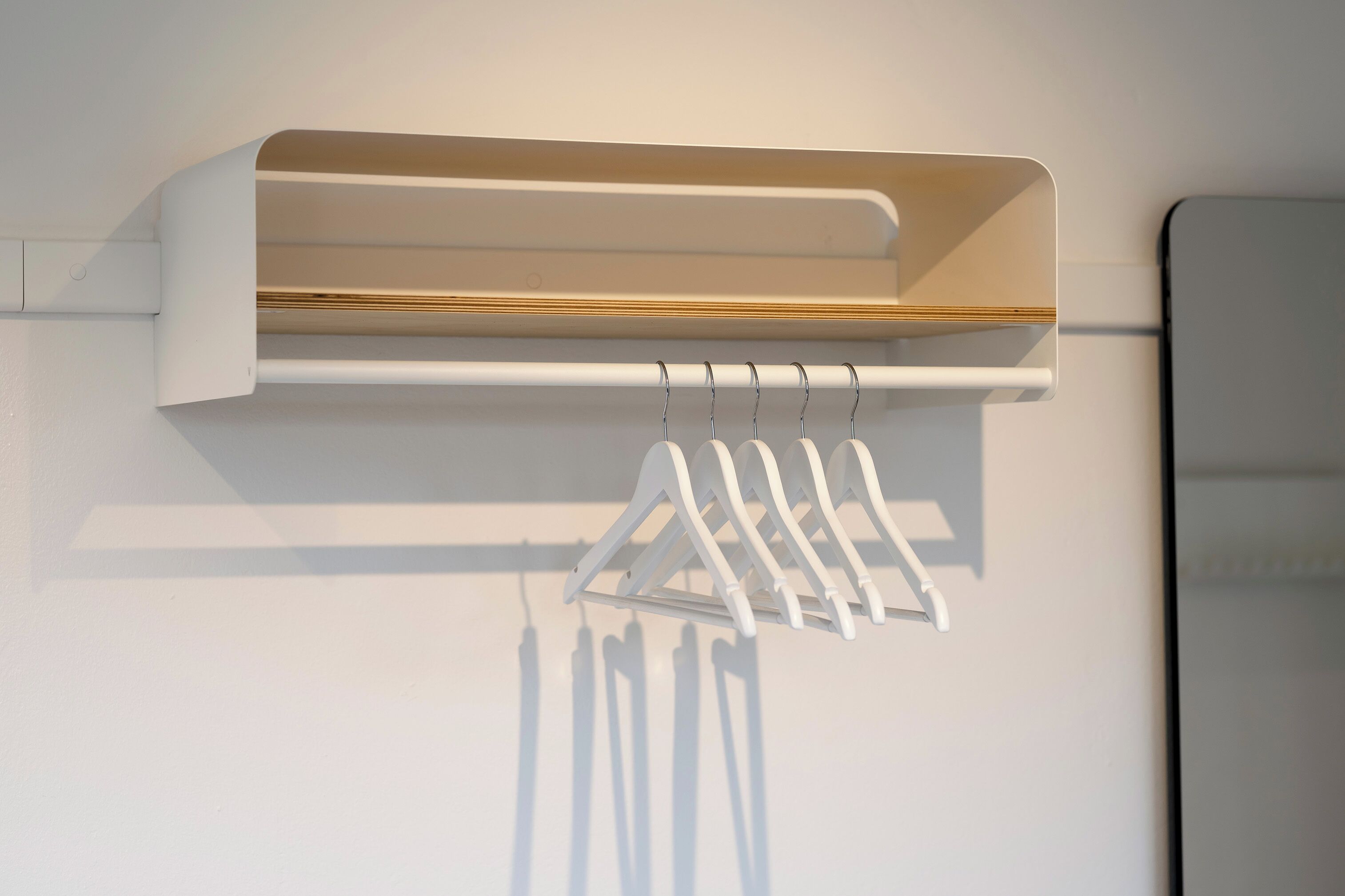 A white wall-mounted shelf with a wooden panel and a white hanging rack with clothes.