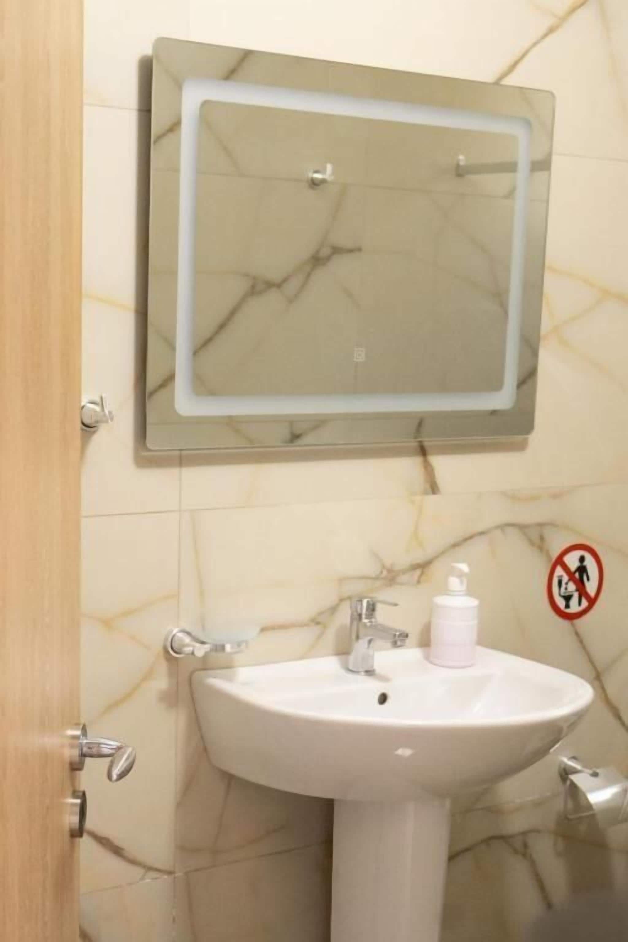 A bathroom with a marble-tiled wall, a white sink with a soap dispenser, and a mirror with a light.