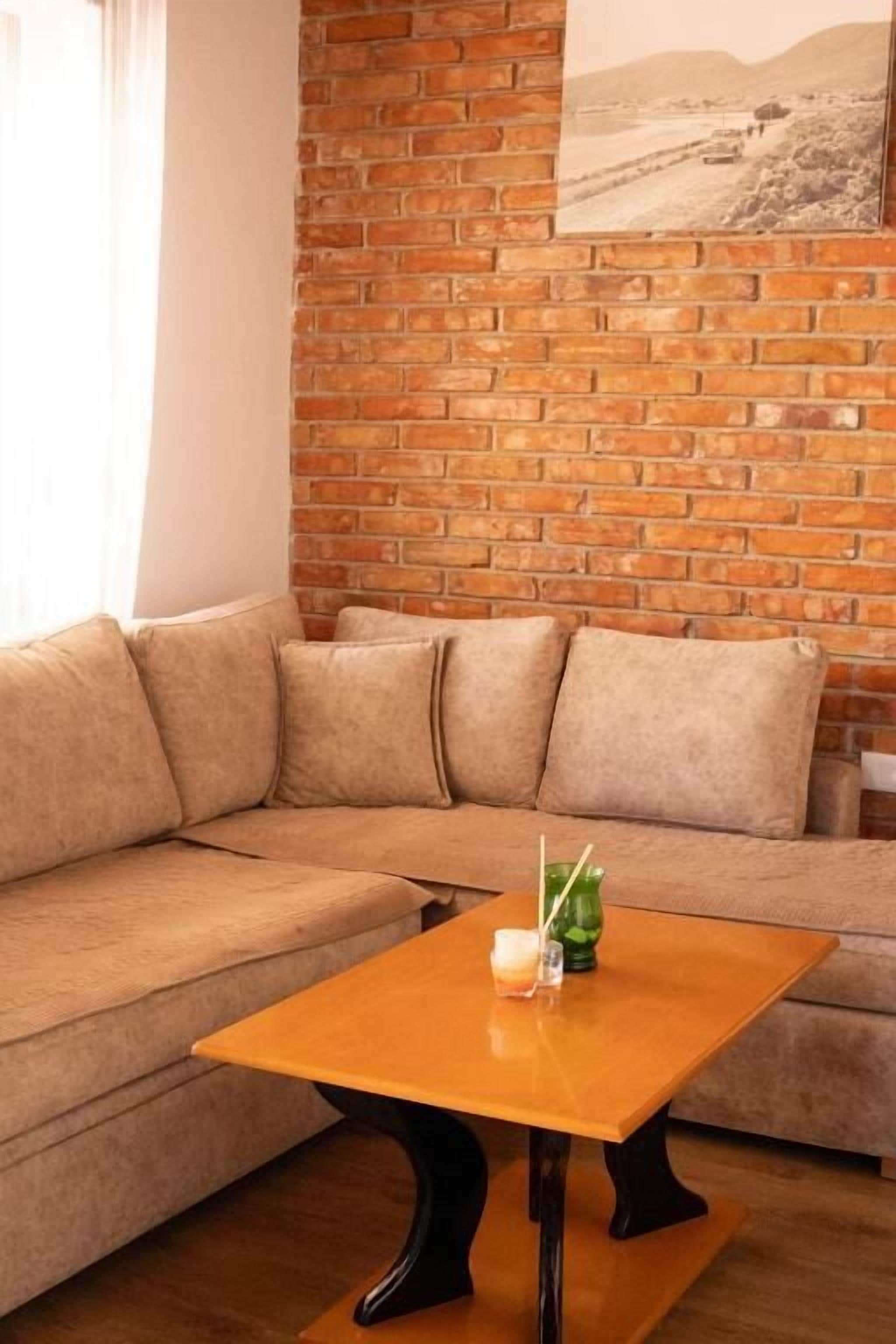 Um quarto com parede de tijolos, um sofá bege e uma mesa de centro de madeira com uma vela e um vaso verde.