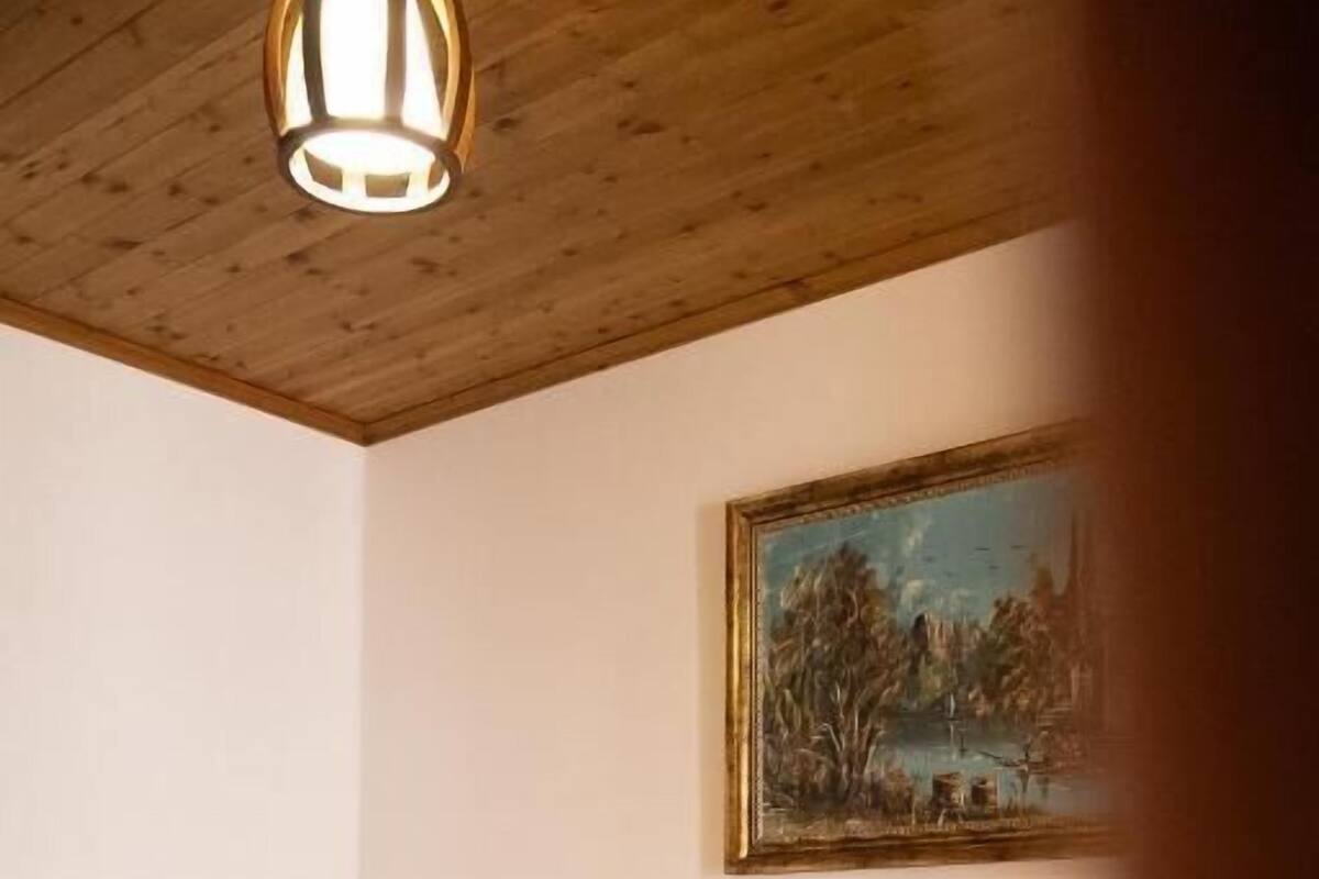 A bedroom with a wooden ceiling, a bed with white linens, a nightstand with a lamp, and a painting on the wall.