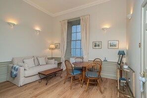 Ein Zimmer mit einer Couch, einem Esstisch mit Stühlen, einer Lampe und einem Fenster mit Vorhängen.
