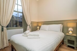 Ein ordentlich gemachtes Bett mit weißen Leinen, ein Fenster mit Vorhängen und zwei Nachttischlampen.