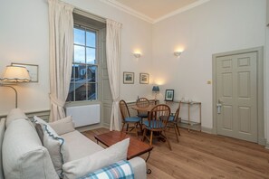 Ein Wohnzimmer mit einer Couch, einem Couchtisch, einem Esstisch mit Stühlen, einem Fenster mit Vorhängen, Wandleuchten und einer Tür.