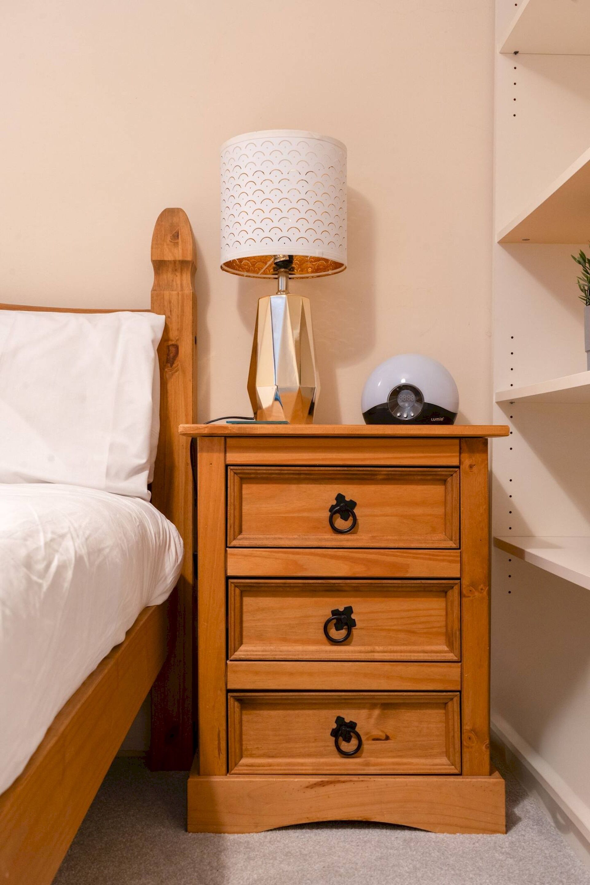 A wooden bedside table with a lamp, a small shelf with a decorative item, and a bed with white bedding.