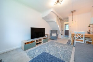 A modern living room with a flat-screen TV, a grey armchair, a wooden dining table, and a staircase leading to another level. - Quiet St. Albans Cottage with Free Parking - Pass the Keys (St Albans)