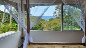 Quarto com uma janela grande, cama com colcha texturizada e vista para vegetação e água.