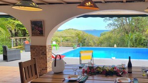 A dining table with chairs, a vase with flowers, and a bottle of wine, overlooking a swimming pool and lush greenery. - Villa Goyave, large family home with sea view and swimming pool! (Le Robert)