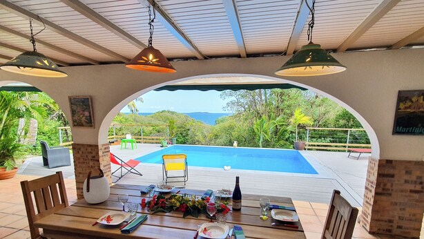 Uma área de refeições ao ar livre coberta com vista para a piscina, móveis de madeira e luminárias pendentes.