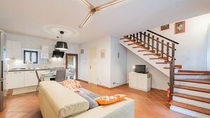 A modern kitchen with white cabinets, a dining area with a table and chairs, a sofa with orange cushions, and a wooden staircase.