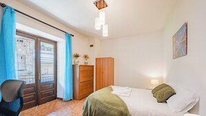 A bedroom with a bed, a wooden wardrobe, a desk with a chair, a window with curtains, and a painting on the wall.