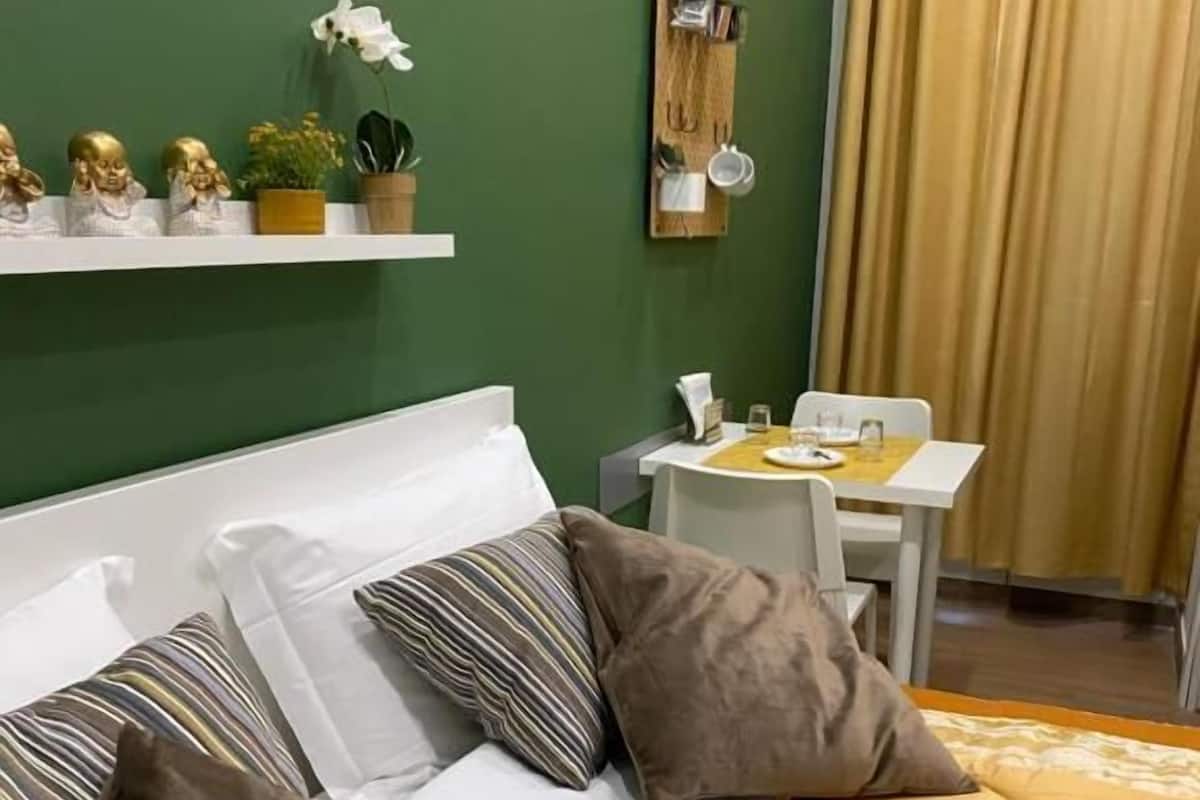A hotel room with a bed, a small table, a chair, a shelf with decorative items, and a curtain.