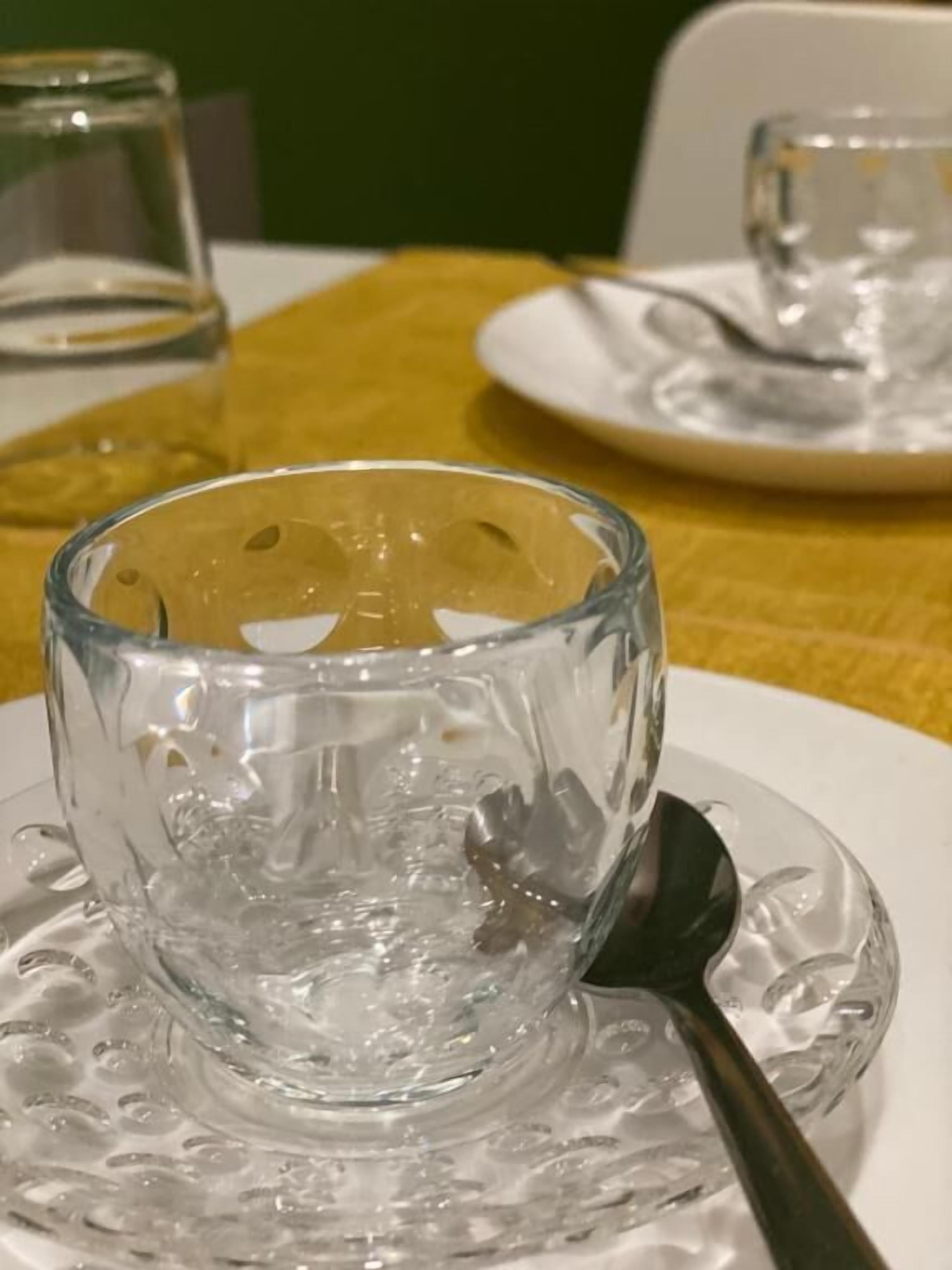 A clear glass cup on a saucer with a spoon, placed on a table with a yellow tablecloth.