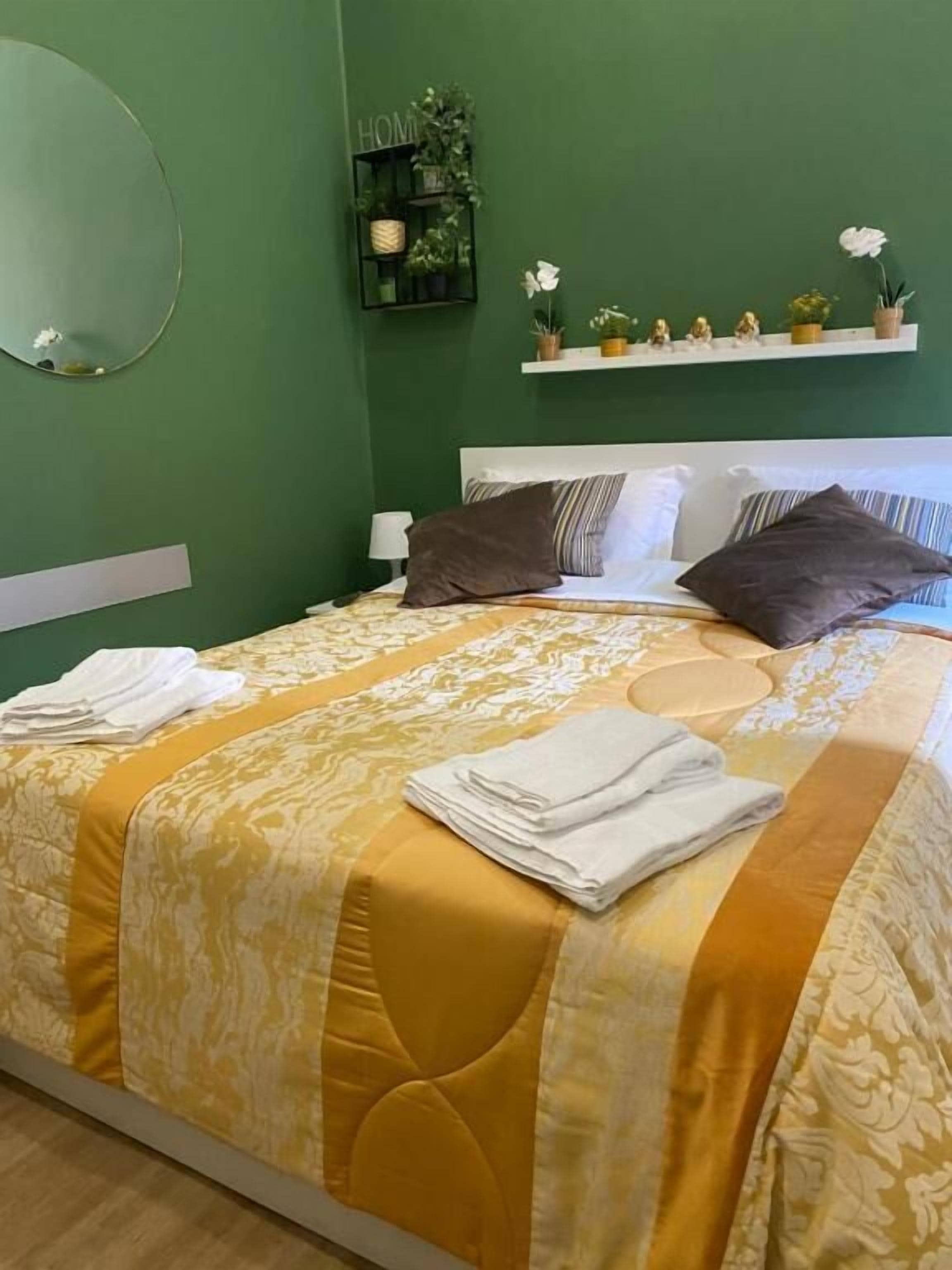 Una cama bien tendida con una colcha amarilla y blanca, un cabecero blanco y una pequeña estantería con plantas.