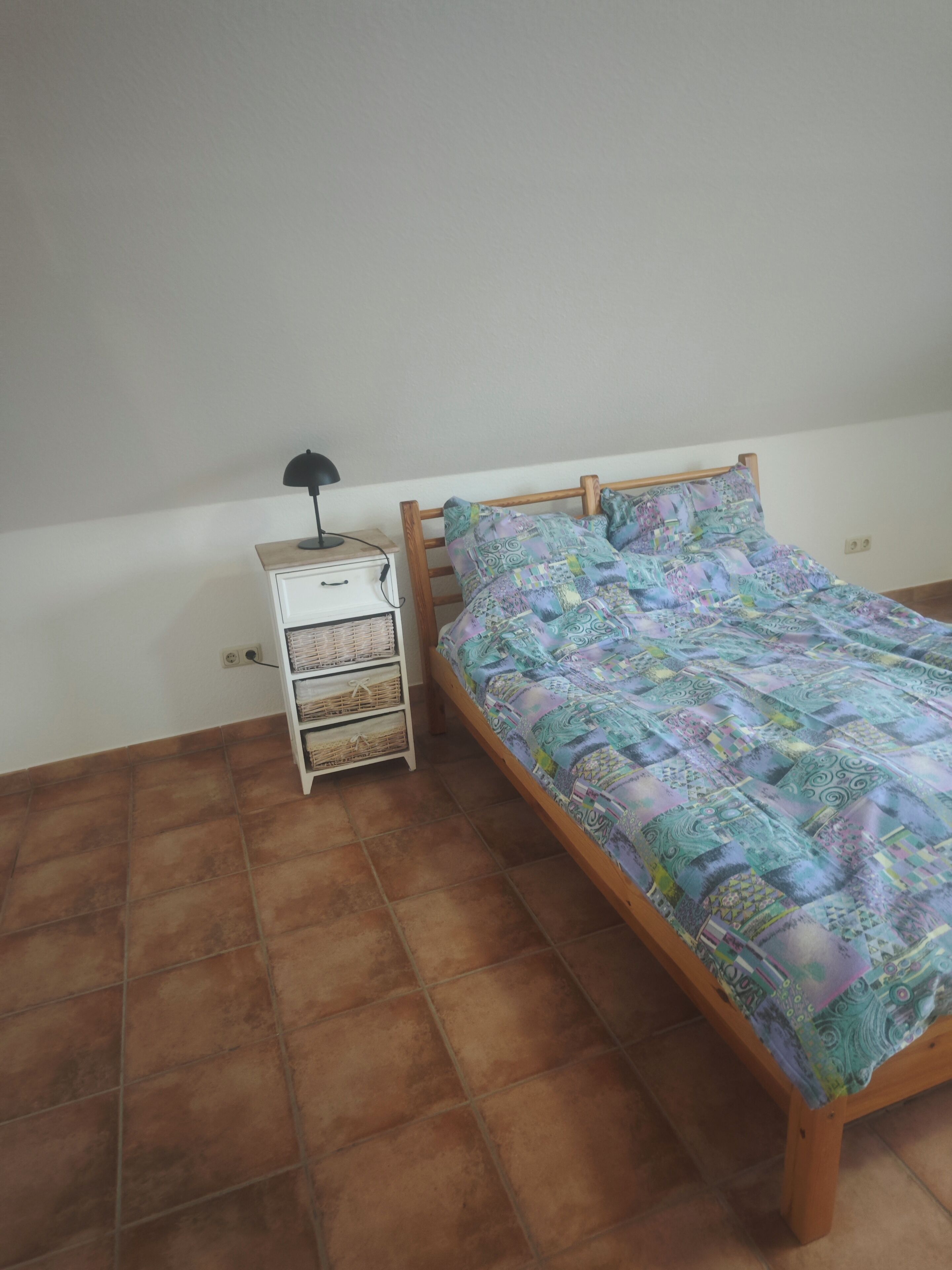 Een eenpersoonsbed met een bedsprei met dessin, een bedlampje en een klein nachtkastje in het wit met lades en manden.