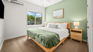 A bedroom with a bed, a nightstand, a ceiling fan, a window with a view, and a door.