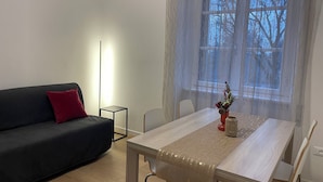 A modern living room with a dark sofa, a small side table, a dining table with a light wood finish, and a window with sheer curtains.