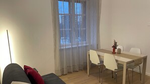 A room with a sofa, a dining table with chairs, a window with sheer curtains, and a lamp.